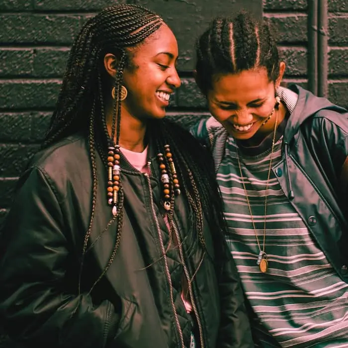 Two friends share a joyful moment, laughing together against a textured brick wall. They are both wearing casual jackets, with one friend having long braided hair adorned with beads, and the other sporting a short, natural hairstyle. Their genuine smiles reflect a deep bond and carefree happiness.