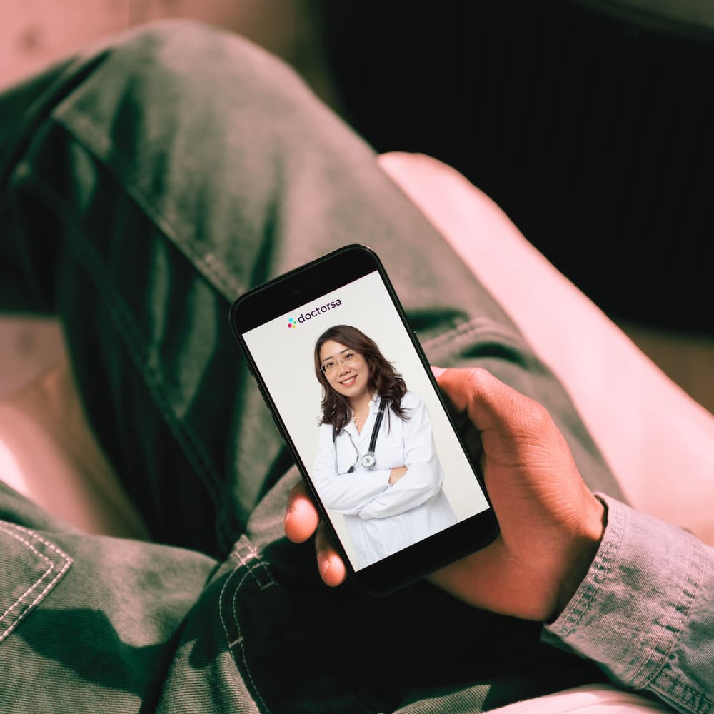A person is holding a smartphone displaying a video consultation using Doctorsa telehealth platform. The patient appears relaxed, dressed casually, possibly engaging in a remote consultation with a UTI doctor in Cancun.