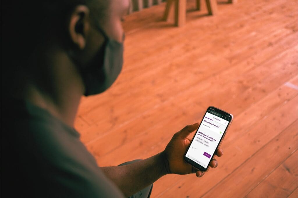 A person with a face mask is holding a smartphone and interacting with Doctorsa. The screen shows options to select a city and choose the type of healthcare service needed. The person is seated and appears to be focused on using the app to find a UTI doctor in Rome.